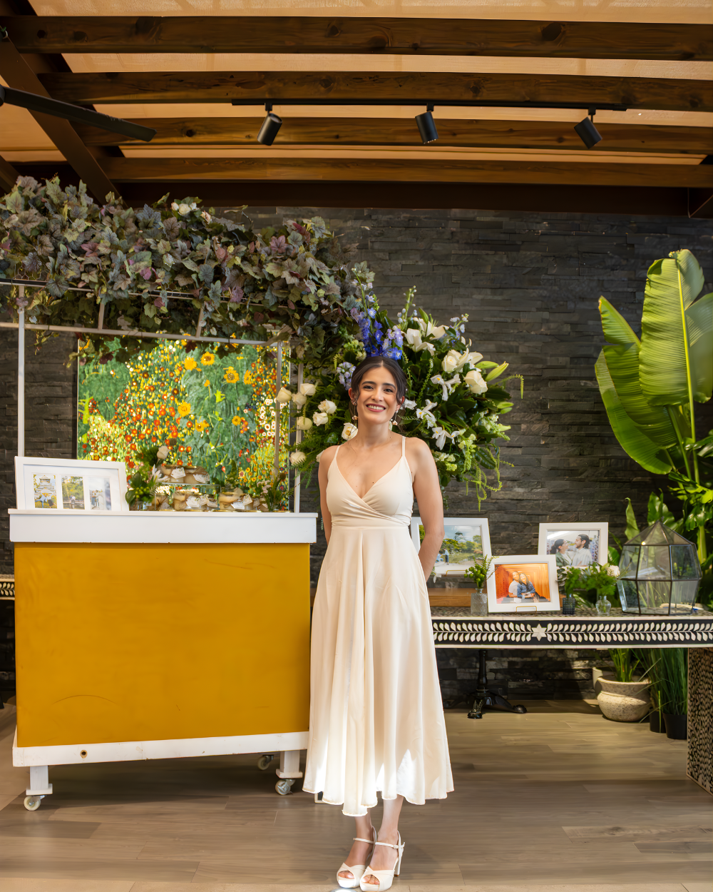 Mujer con vestido color crema de pie junto a una exhibición floral para eventos con mostrador amarillo y fondo de vegetación.