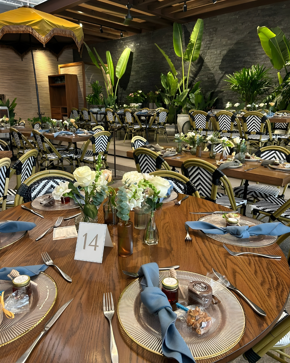 Mesa del salón de banquetes puesta con platos a rayas, servilletas azules, flores y el número 14 bajo plantas tropicales.