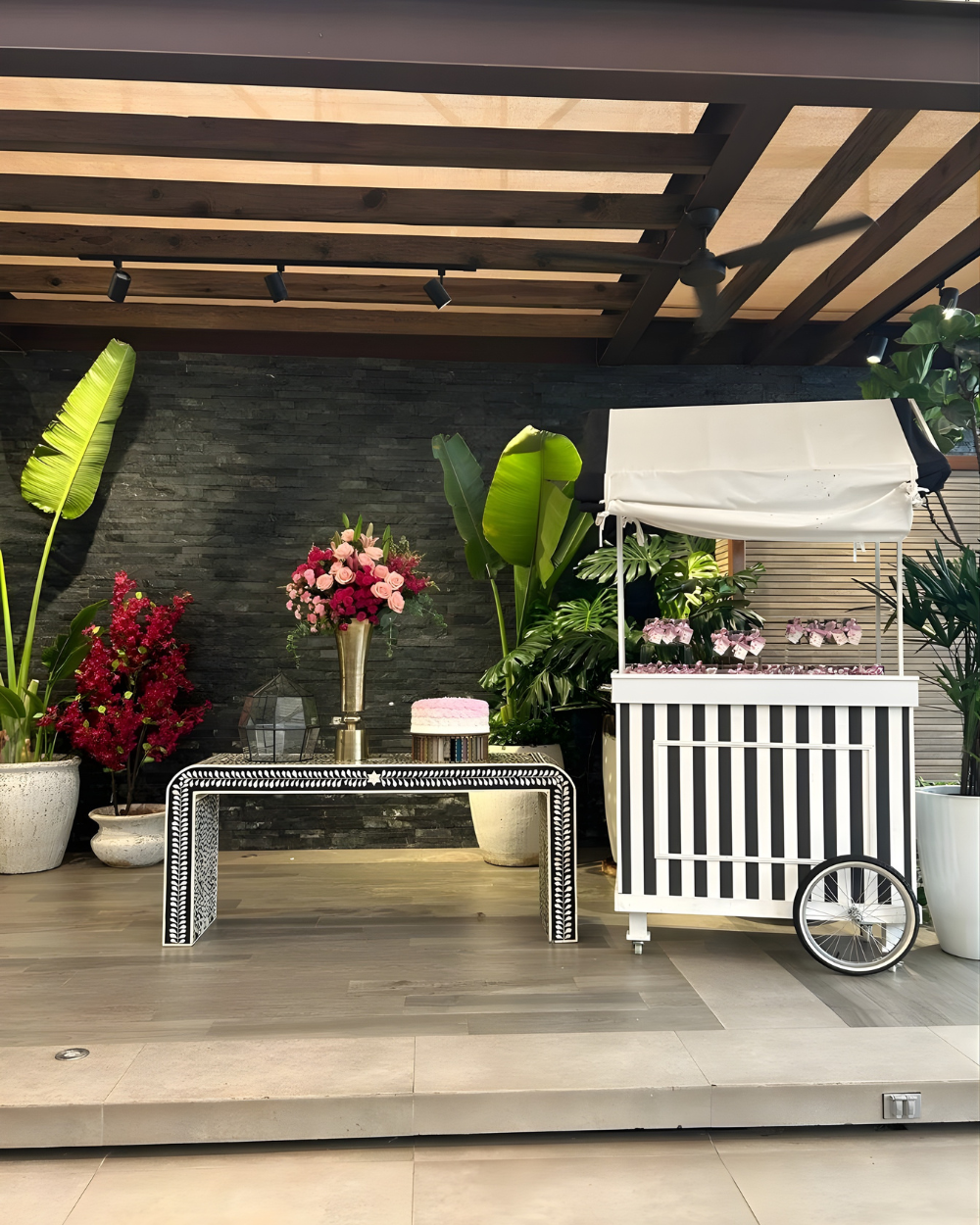 Puesto de mercado al aire libre con toldo blanco, macetas con flores y mesa decorativa bajo una pérgola de madera.