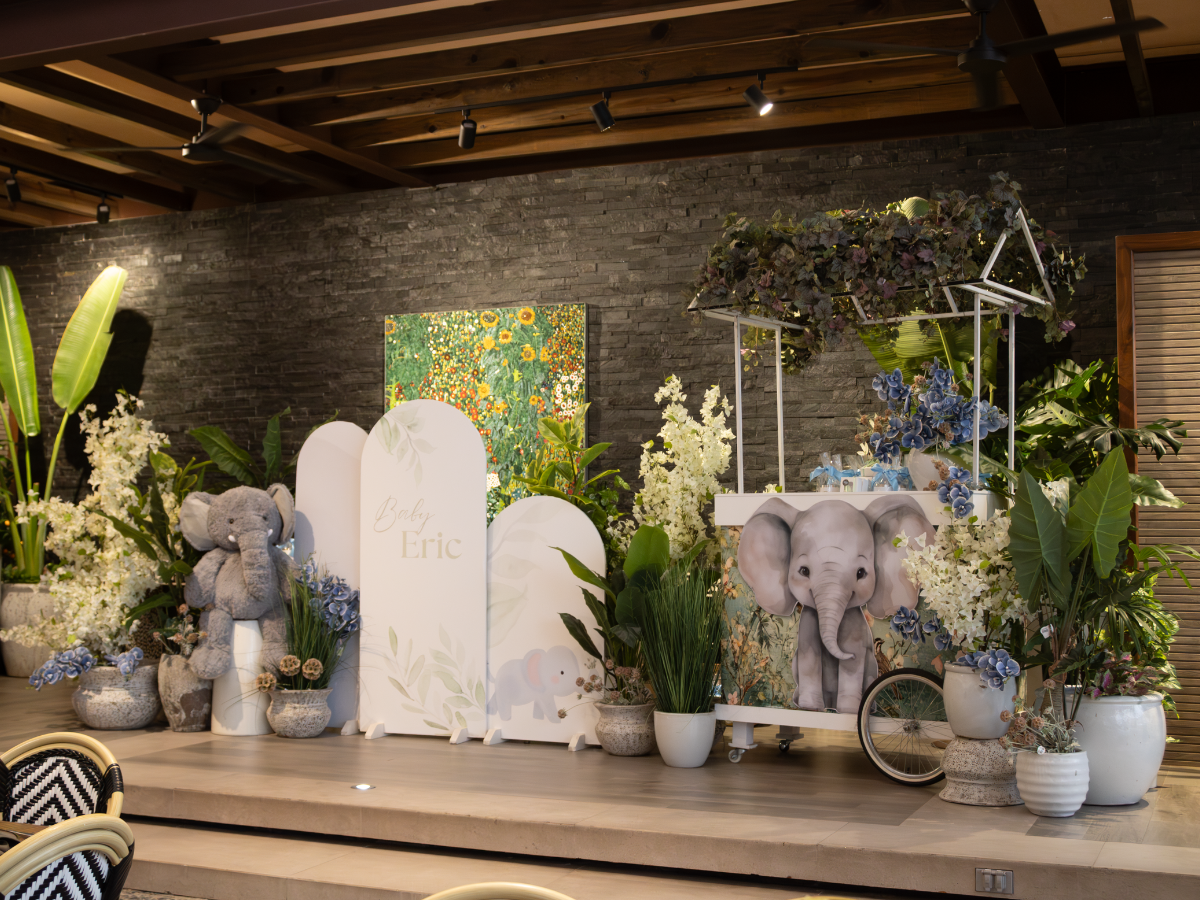 Exhibición decorativa floral y de elefantes sobre un escenario con plantas, paneles blancos y un colorido tapiz de pared.