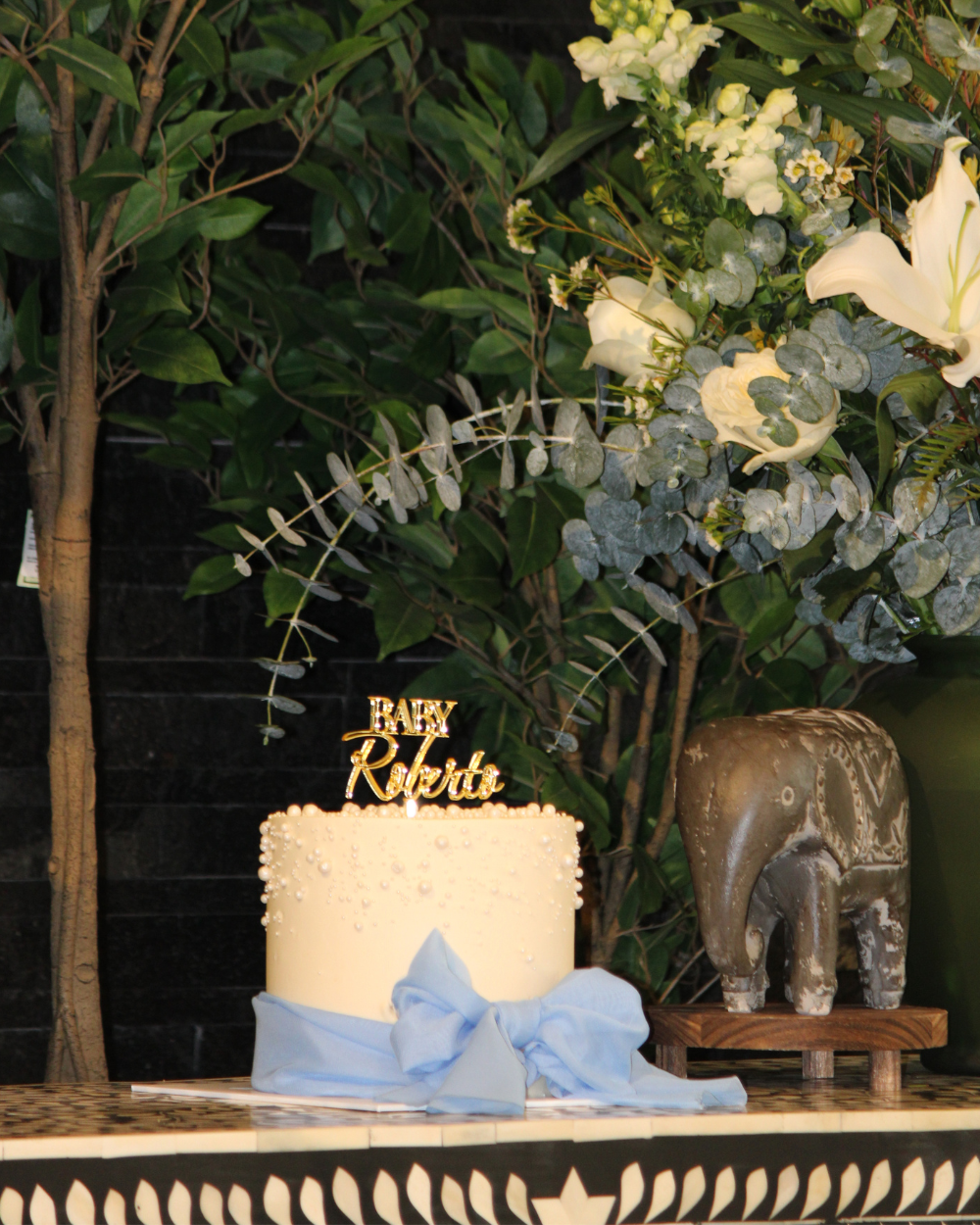Tarta de cumpleaños con adorno dorado y cinta azul sobre una mesa decorada junto a una figurita de elefante y flores.