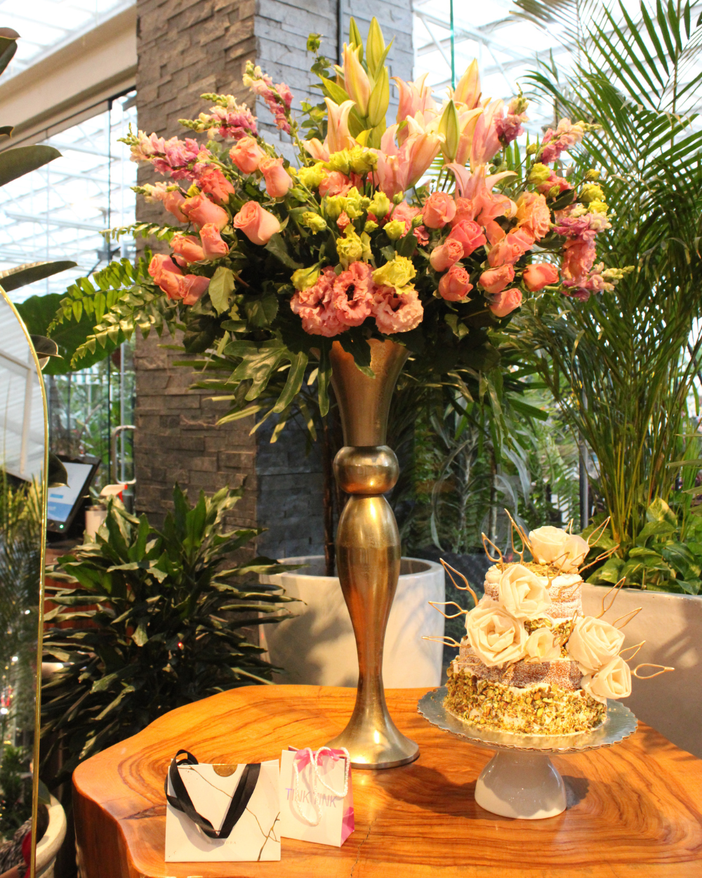 Elegante arreglo floral en un jarrón dorado sobre una mesa de madera junto a un pastel de varios pisos y una caja de regalo.