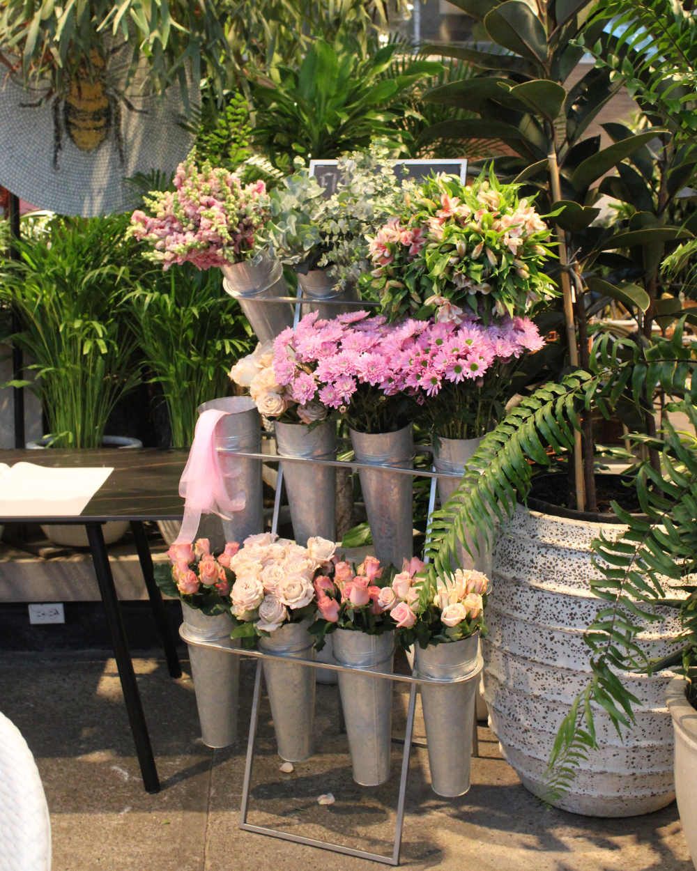 Soporte metálico escalonado con macetas de flores rosas, melocotón y verdes en un entorno de tienda de jardinería.