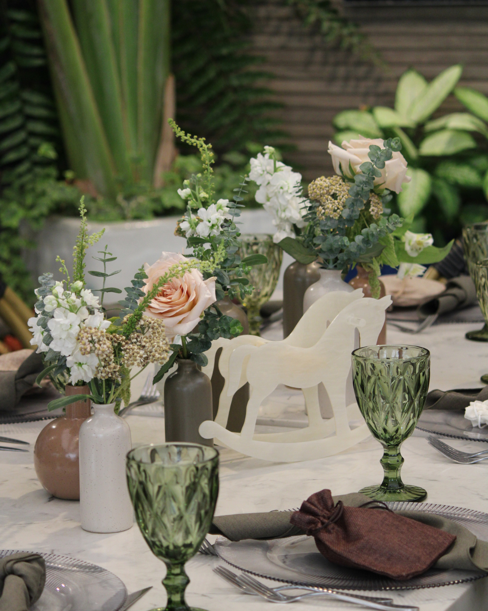 Elegante mesa de exterior decorada con flores en tonos pastel, copas verdes y figuritas decorativas blancas.