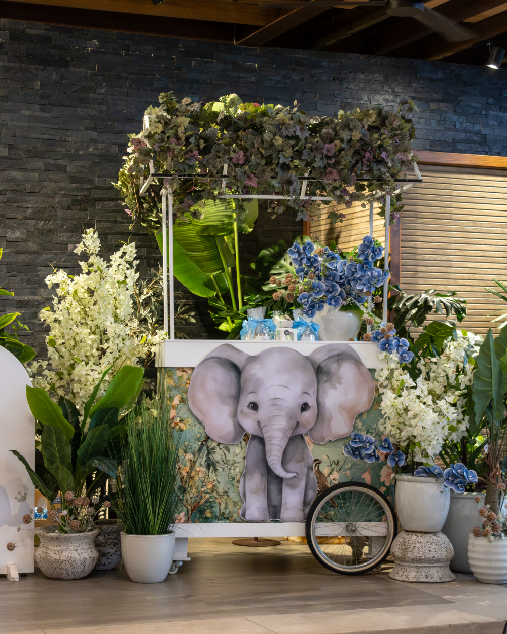 Centro de mesa con retrato de elefante rodeado de flores blancas y vegetación en una exhibición de eventos en interiores.