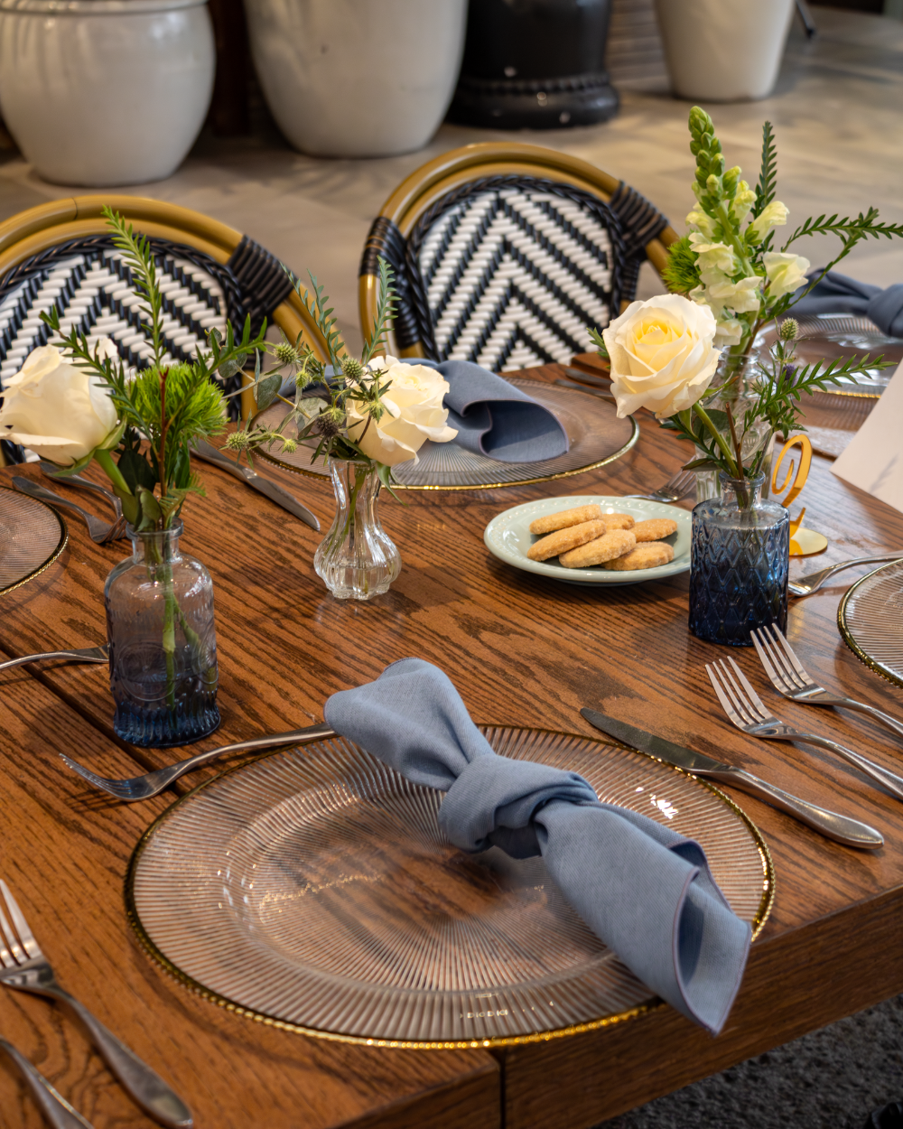 Prepara la mesa del comedor con sillas de mimbre, servilletas azules, flores blancas y cristalería sobre una mesa de madera.
