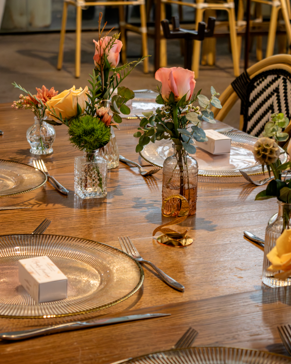 Elegante juego de mesa de comedor con bajoplatos dorados, cajas blancas y pequeños arreglos florales rosas.