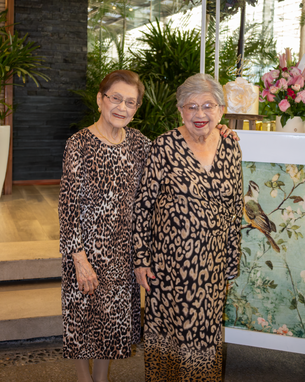 Dos mujeres con vestidos estampados de leopardo están de pie en el interior, cerca de unas escaleras y flores.