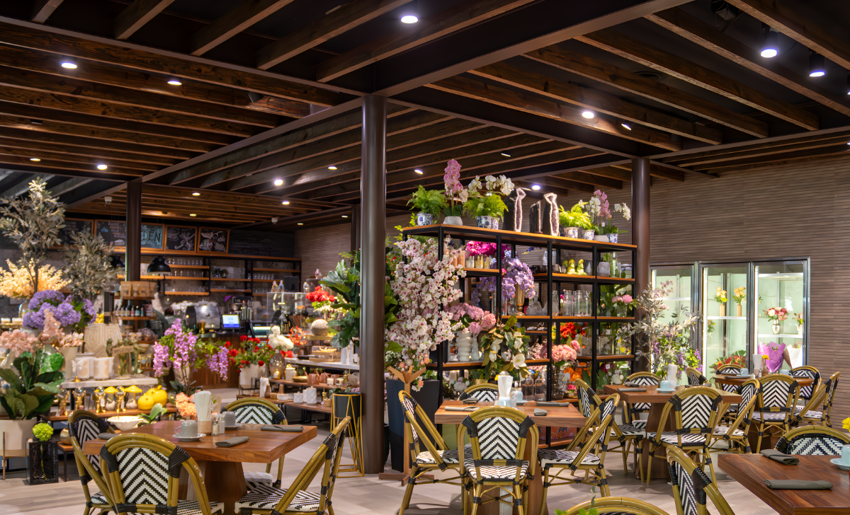 Cafetería luminosa con floristería, mesas de madera, arreglos florales y un techo iluminado con guirnaldas de luces cálidas.