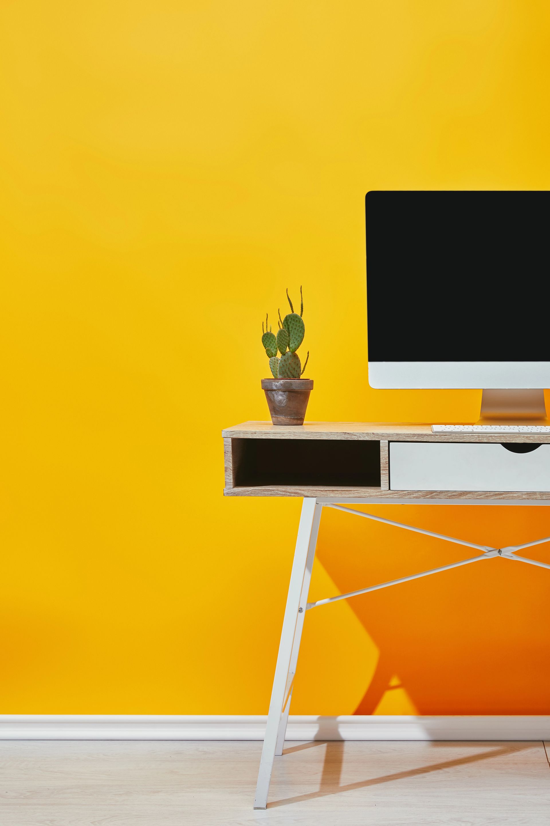 Scrivania minimalista con monitor del computer e pianta in vaso davanti a una parete giallo brillante