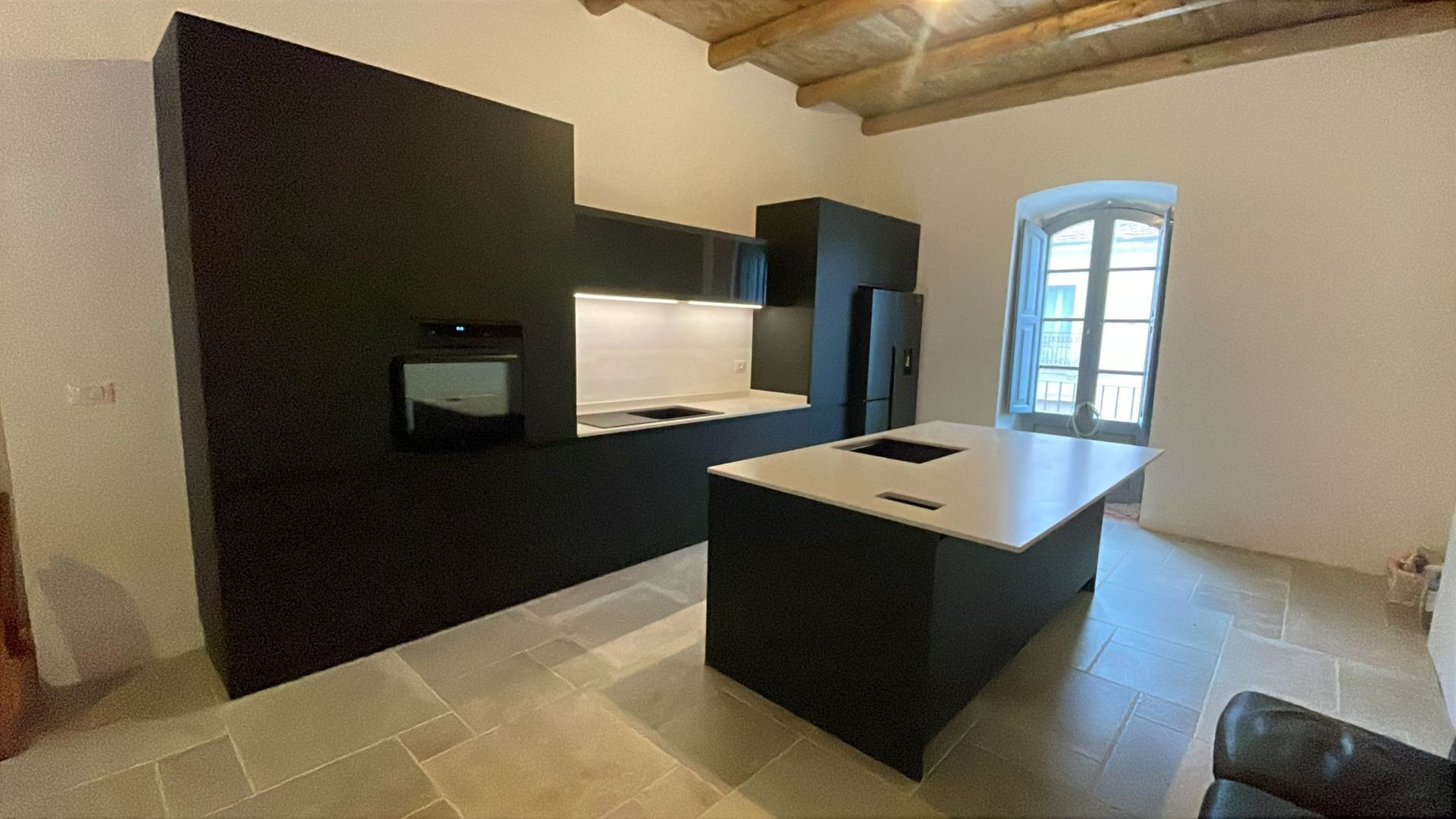 Cucina moderna con mobili scuri, isola centrale, travi a vista e una finestra luminosa.