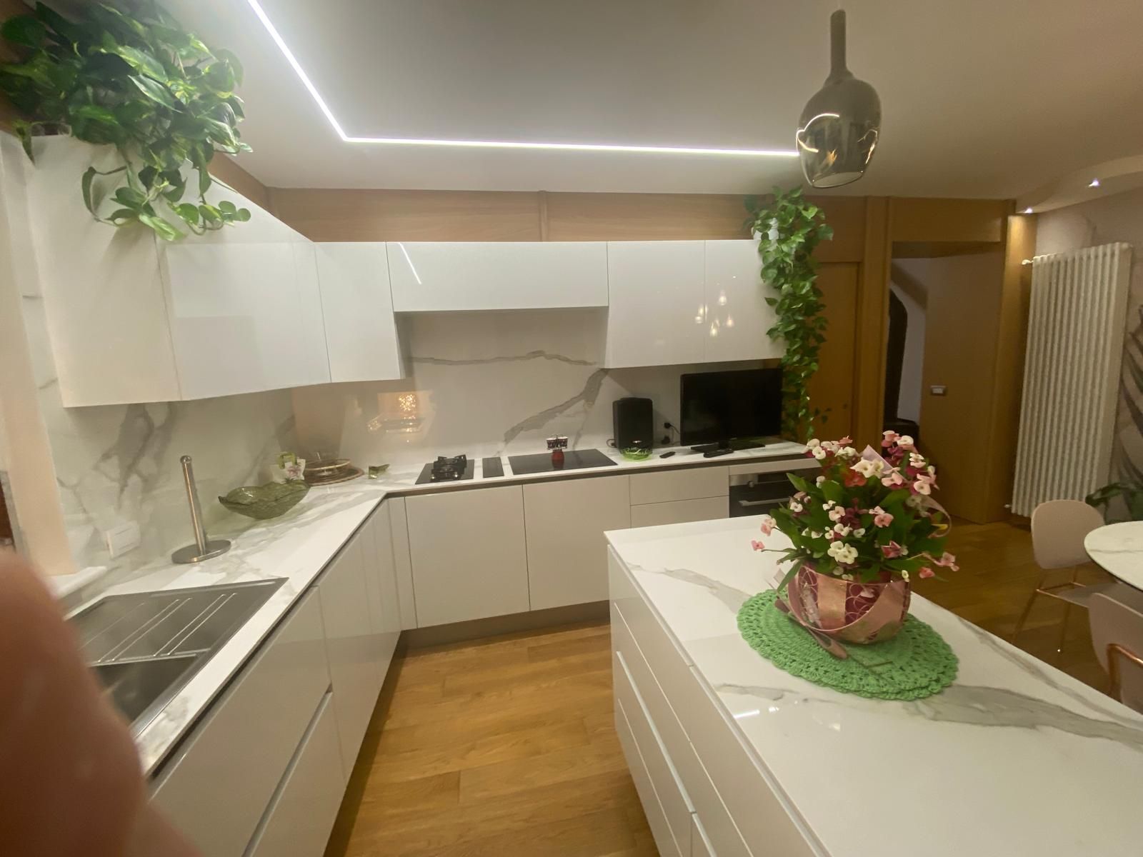 Cucina moderna bianca con pavimento in legno, isola centrale e fiori rosa in un vaso