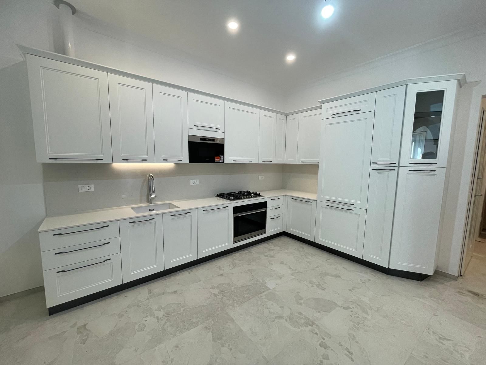 Cucina moderna e luminosa, con mobili bianchi, piani di lavoro grigi, fornelli, lavello e frigorifero a incasso