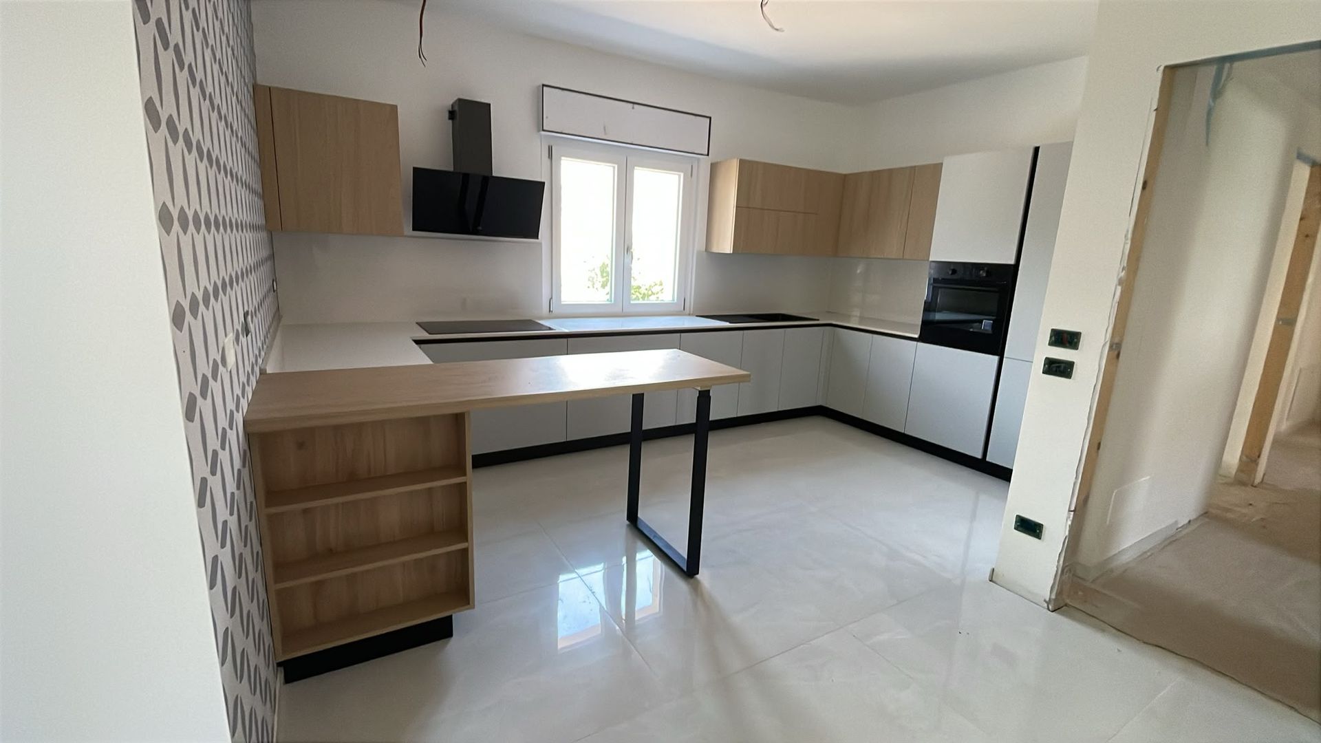 Cucina moderna, luminosa e spaziosa, con mobili bianchi, dettagli in legno, piani di lavoro neri e un’isola centrale.