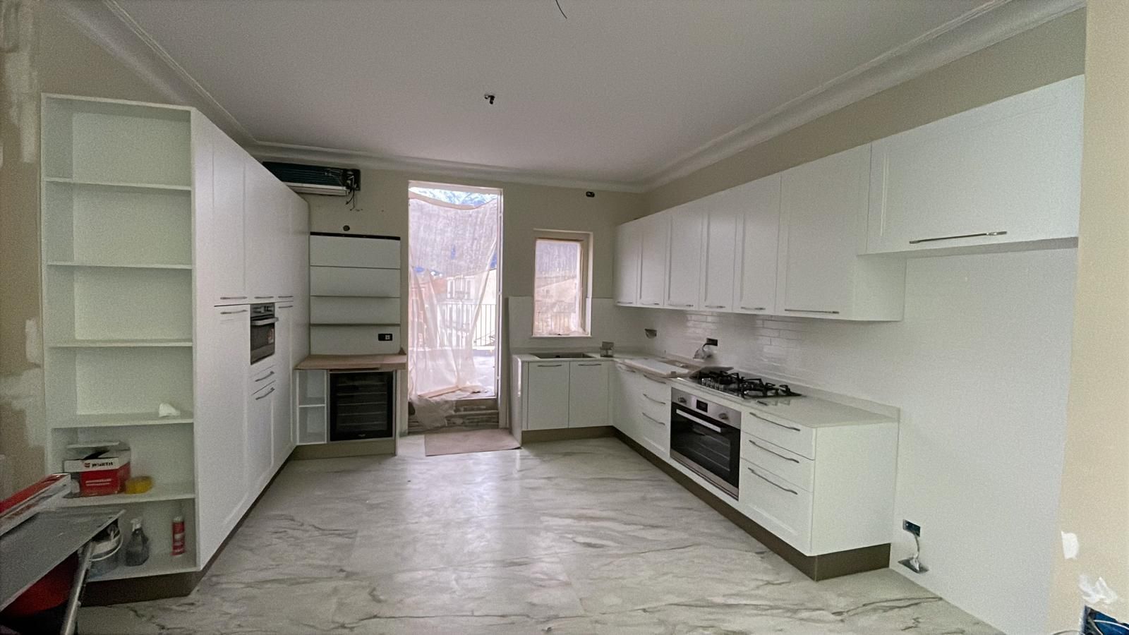 Cucina moderna e luminosa, con mobili bianchi, fornelli, pavimento piastrellato e una porta che dà sull’esterno.