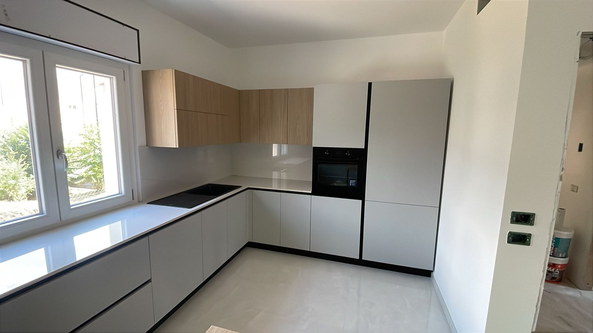 Cucina moderna e spoglia con armadietti bianchi, piani di lavoro neri, dettagli in legno e ampie finestre