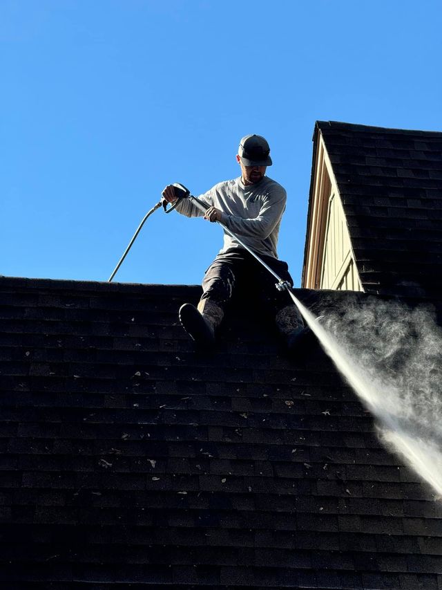 Person power washing a dark shingled roof under a clear blue sky.