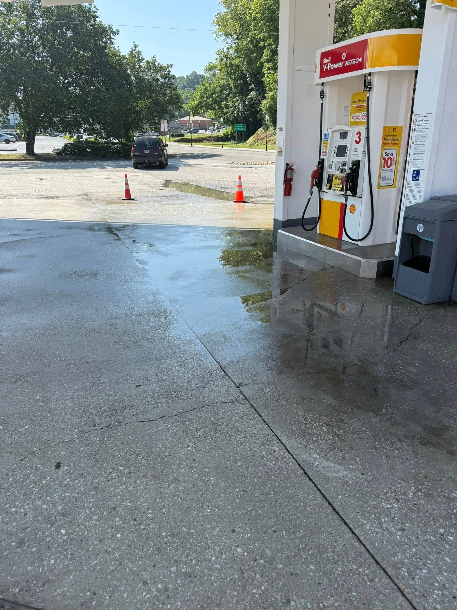 Wet gas station pavement with Shell pumps; orange cones; a vehicle in the distance.