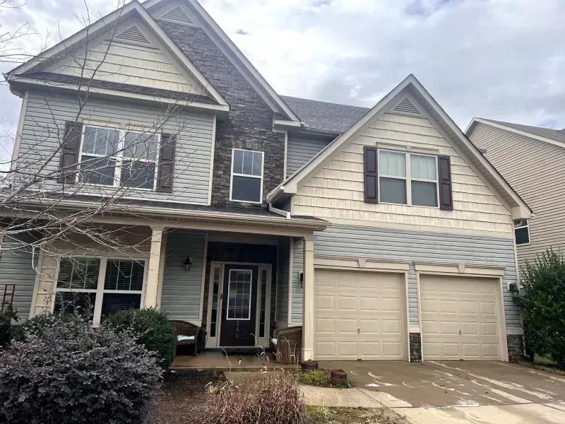 Two-story house with blue siding, stone accents, and tan garage doors under a cloudy sky.