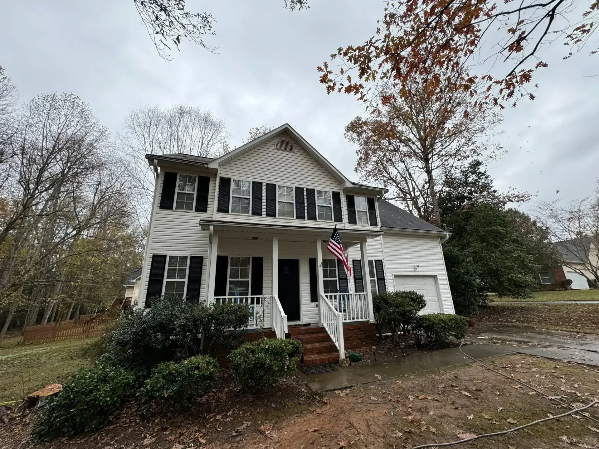 Two-story white house with black shutters, front porch, American flag, and overcast sky.