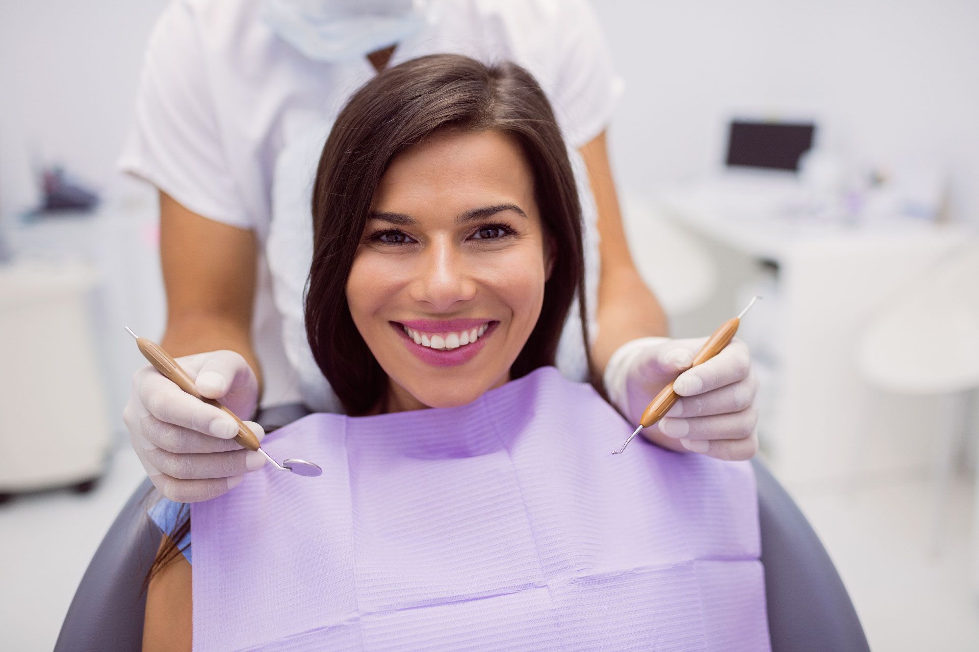 Paciente sonriente en el sillón dental con babero morado mientras el dentista prepara sus herramientas.