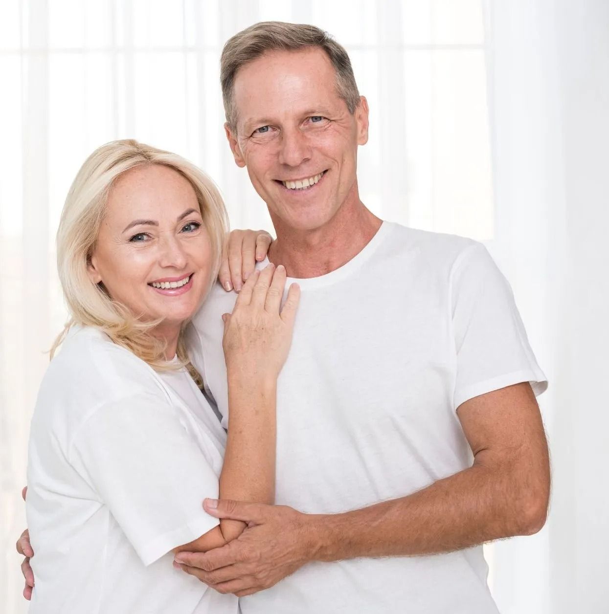 Pareja sonriente con camisas blancas posando muy cerca una frente a un fondo brillante.