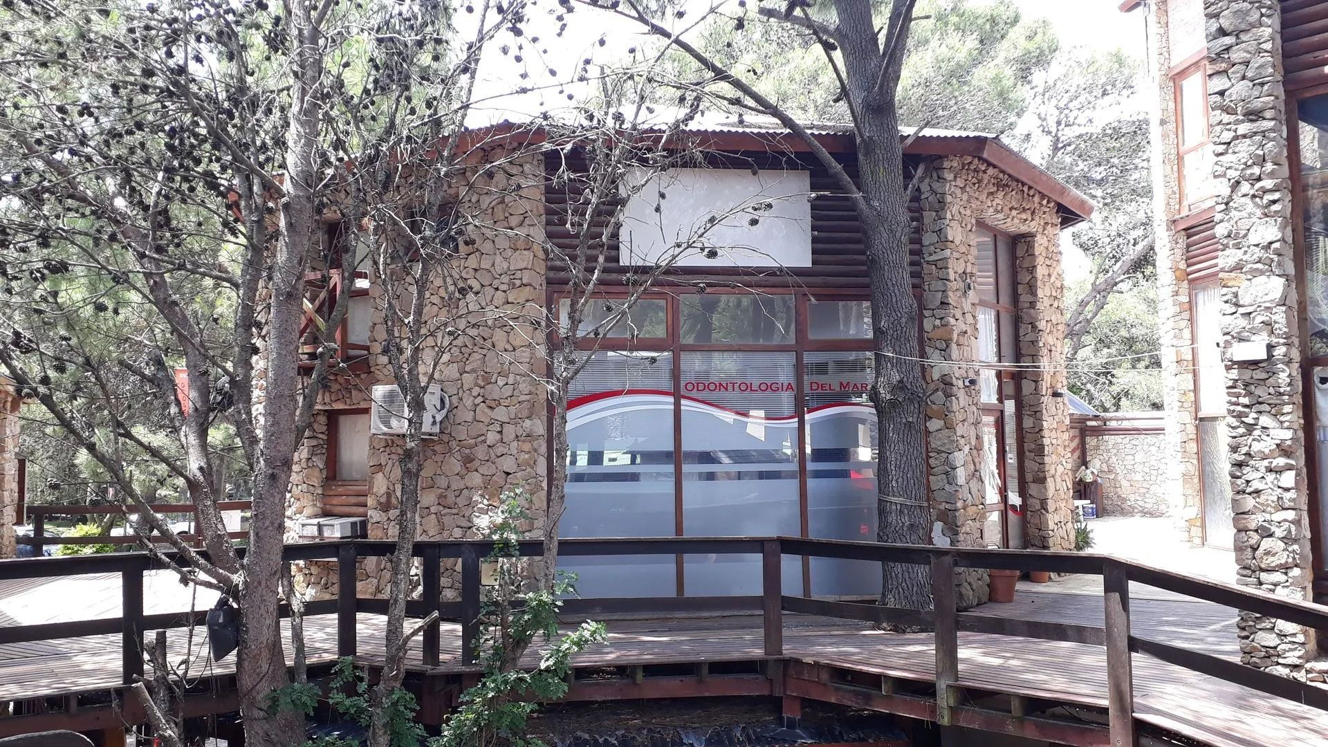 Edificio rústico de piedra con grandes ventanales, visible a través de los árboles y una valla de madera.