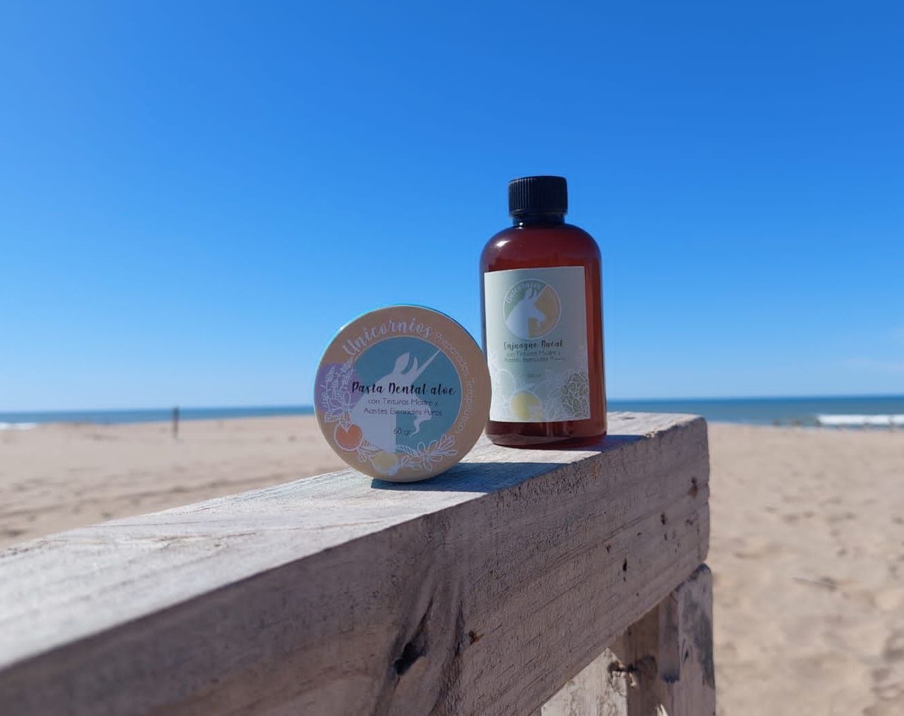 Una botella de color ámbar y un recipiente redondo sobre una barandilla de madera en la playa, bajo un cielo azul despejado.