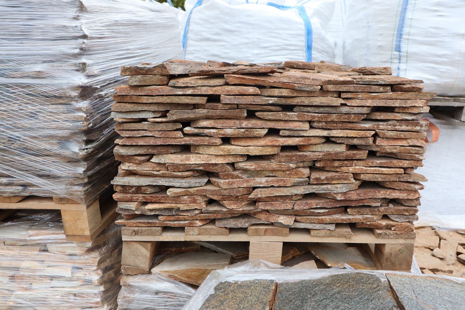 Stacked rough wooden planks on pallets in a storage yard, wrapped materials in the background