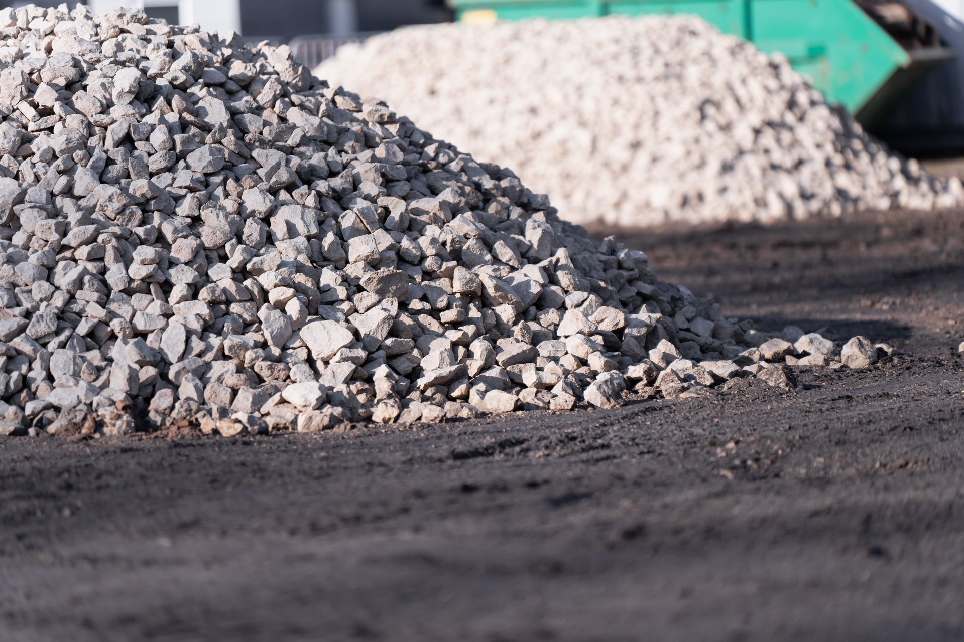 Piles of gravel at a construction site on a dirt surface, with a blurred truck in the background