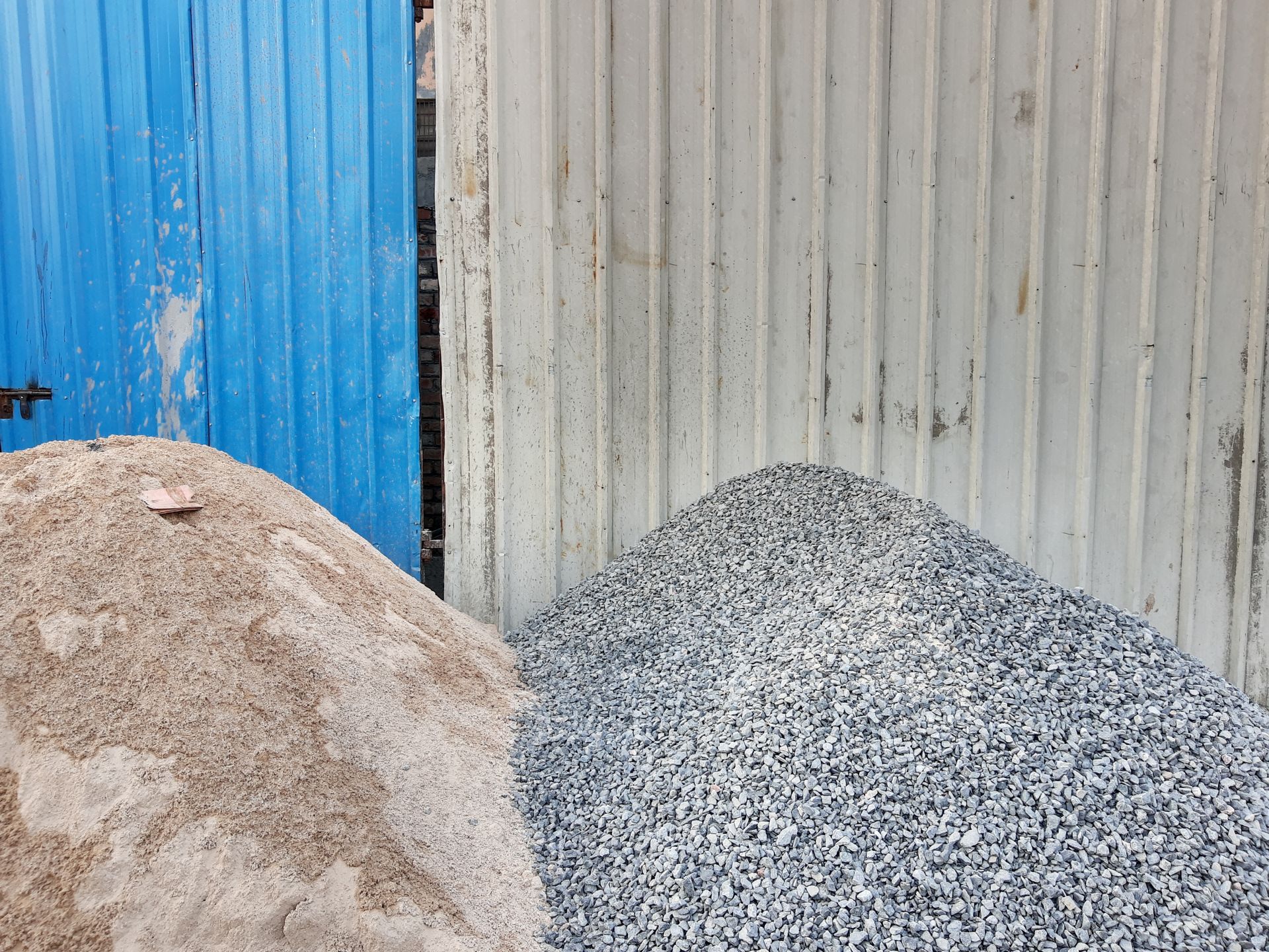 Pile of sand and pile of gravel stored side by side against a corrugated metal wall. Pile of sand and pile of gravel stored side by side against a corrugated metal wall.