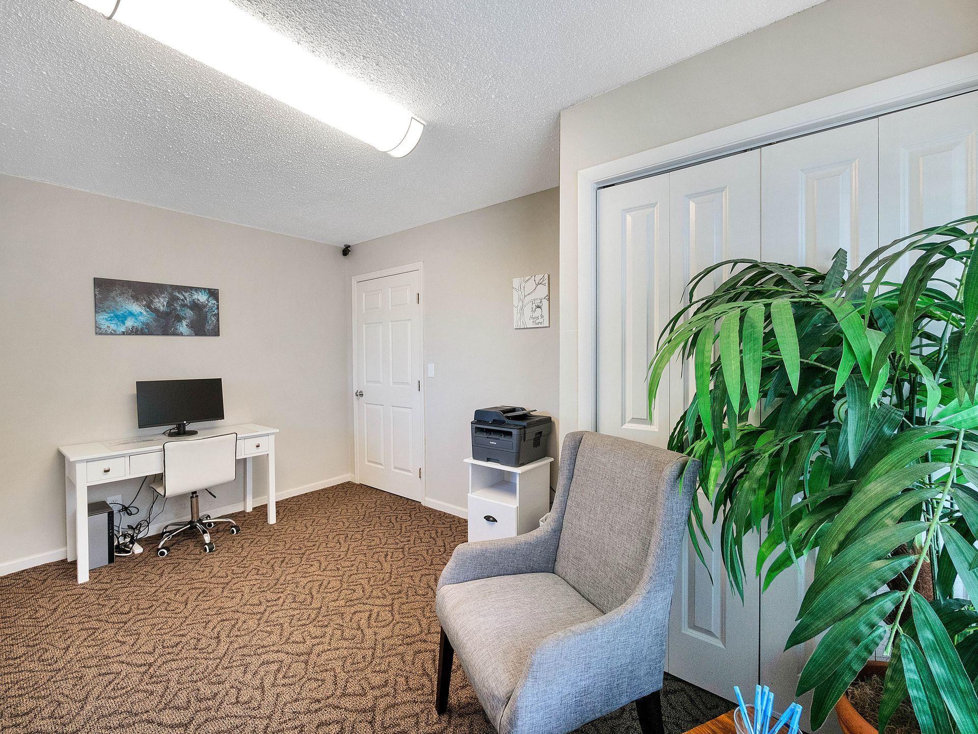 A home office with a white desk, computer, grey armchair, and a large plant in the corner on brown patterned carpet.