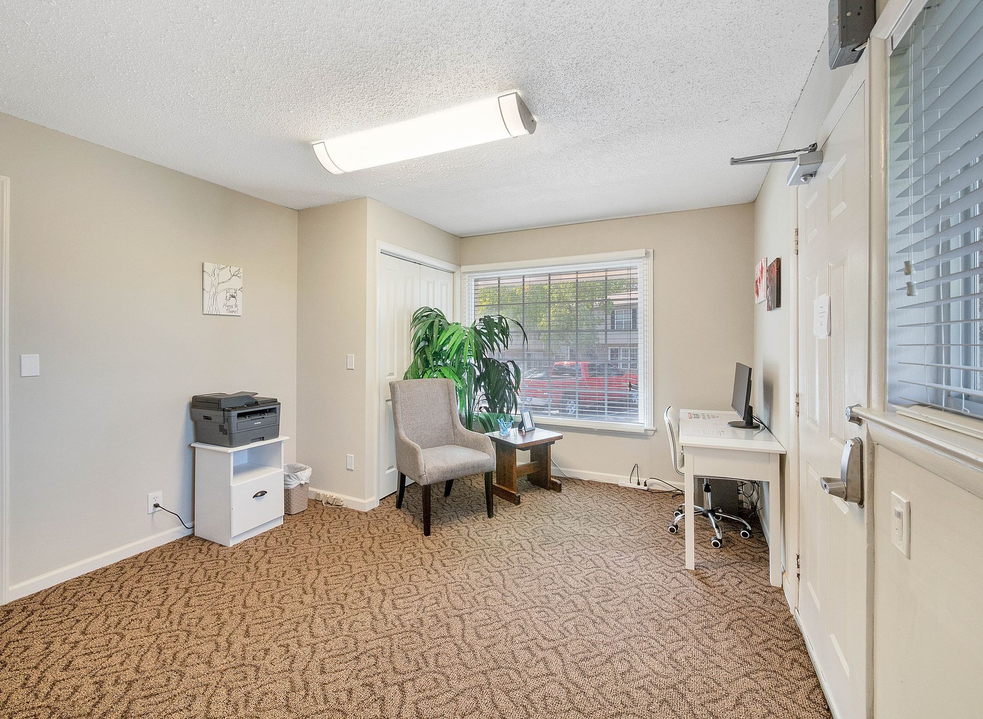 A beige room with patterned carpet, a gray armchair, a printer on a white stand, a desk with a monitor, and a window.