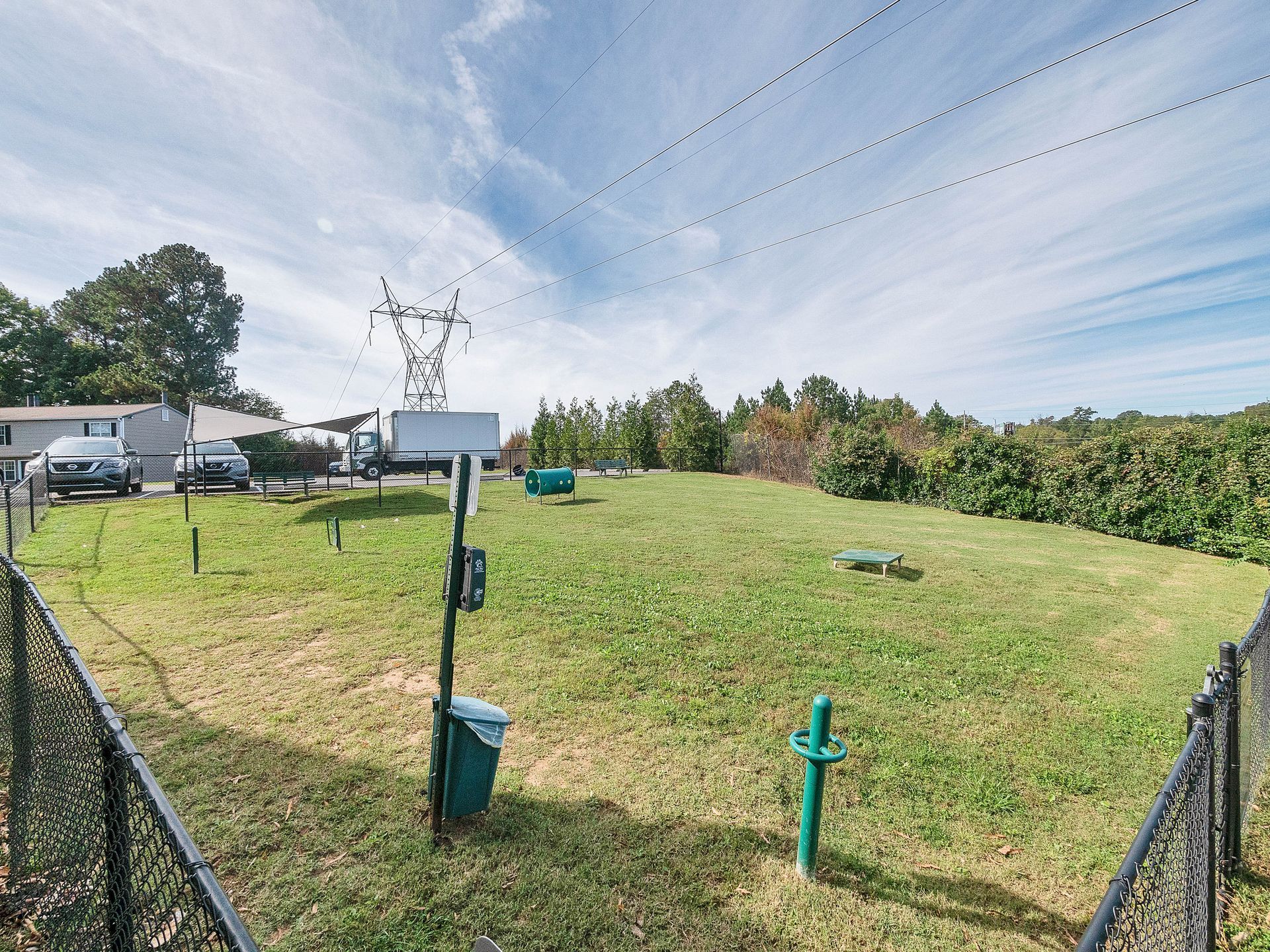 A fenced, grassy lot with utility equipment, power lines overhead, and cars parked in the background under a blue sky.