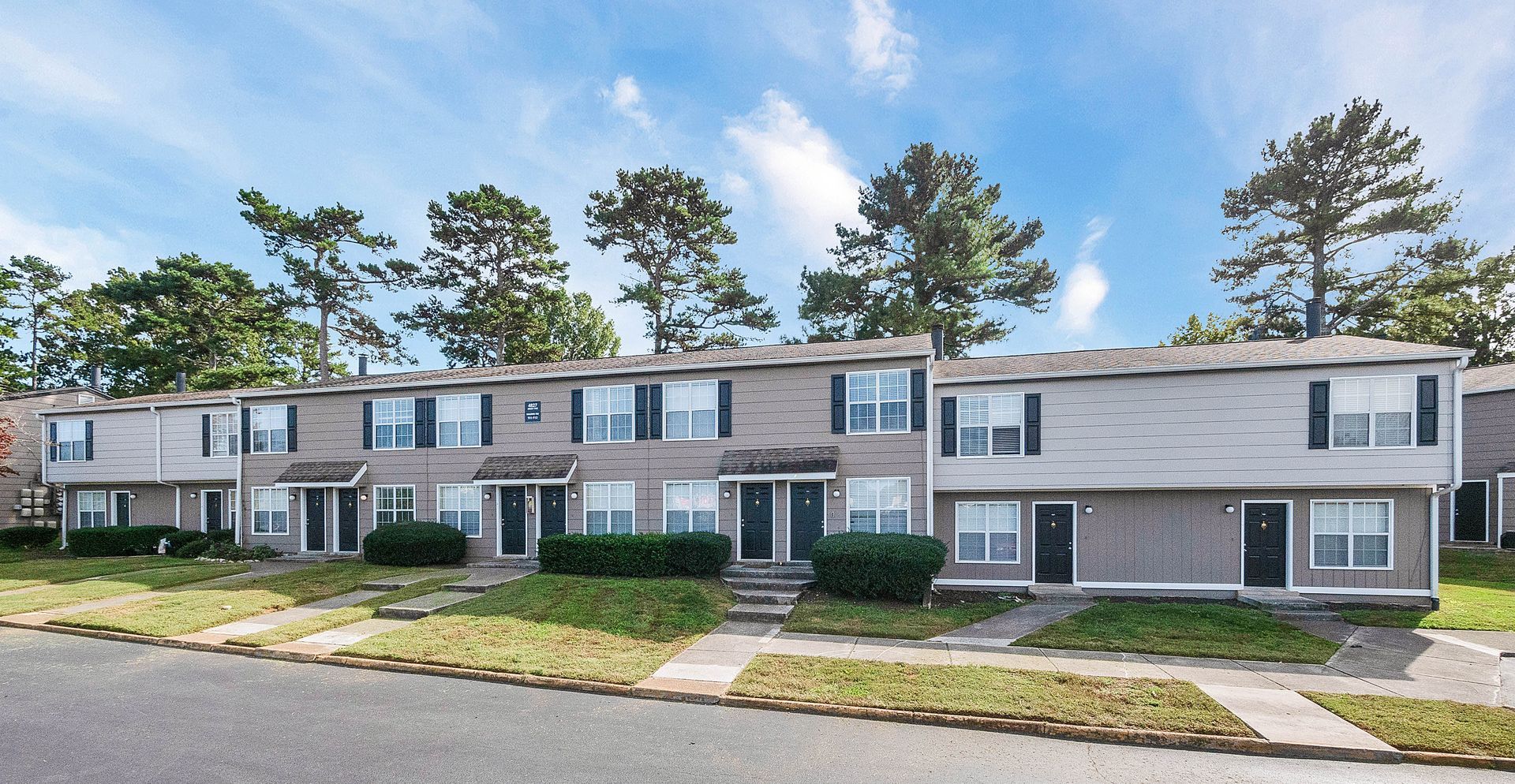 A row of two-story townhouses with light-colored siding, dark shutters, and a front lawn under a blue sky.