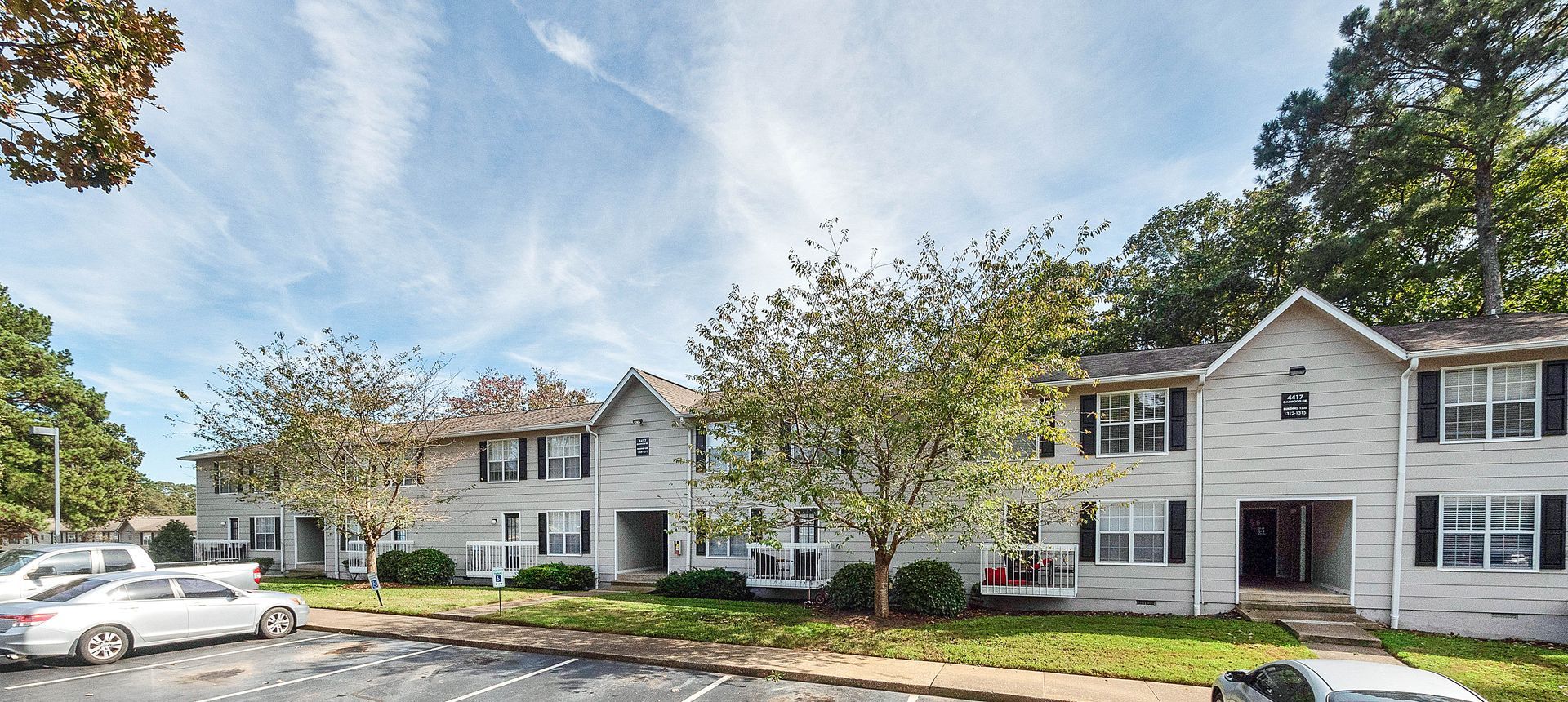 A two-story apartment building with light-colored siding, dark shutters, and a parking lot in front under a sunny sky.