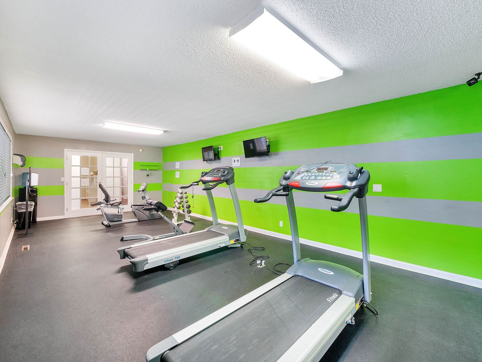 Two treadmills face a bright green and gray striped wall in a fitness room with rubber flooring and overhead lighting.