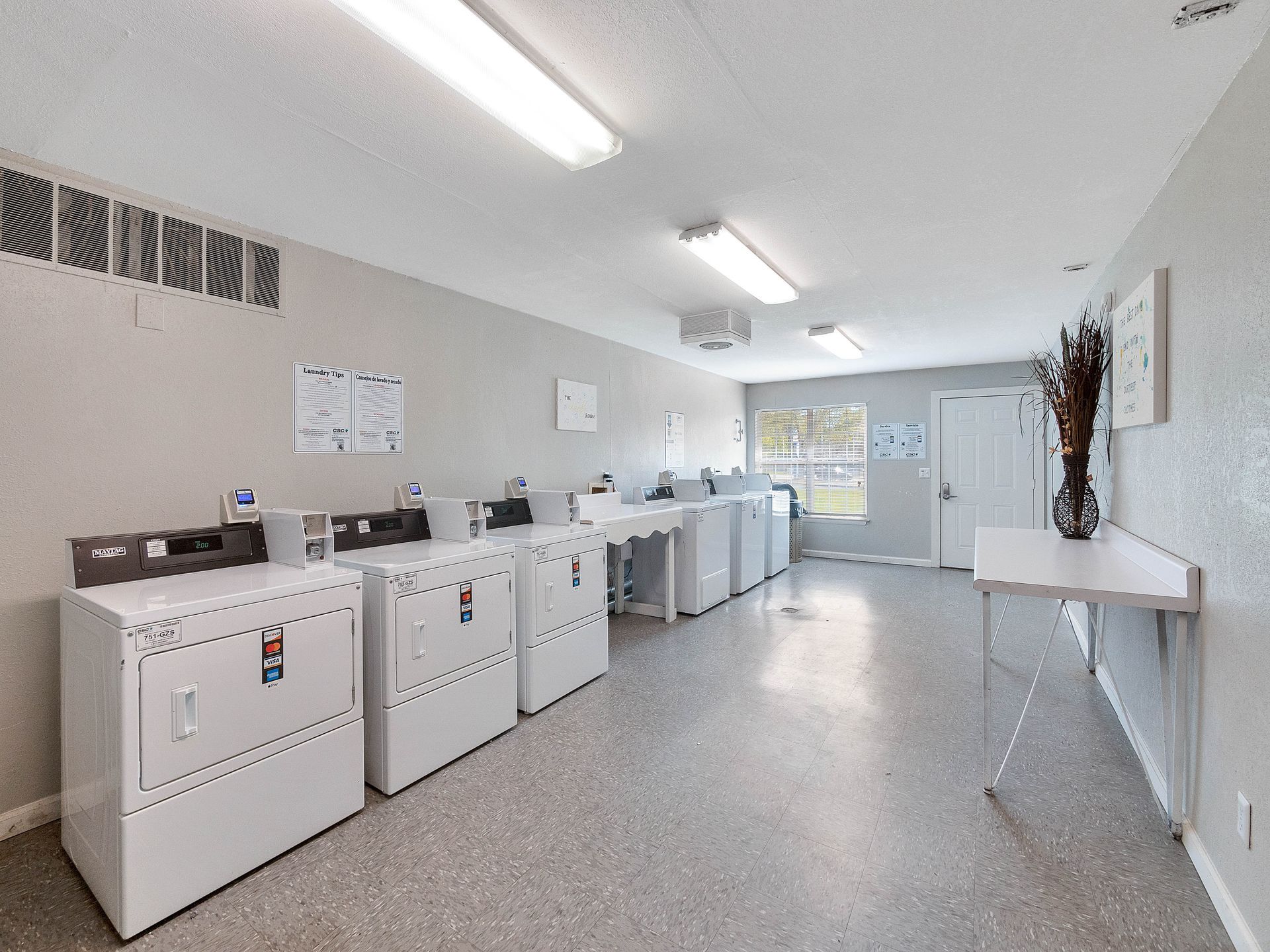 A clean, well-lit laundry room with a row of white dryers, a folding table, and a door at the far end.