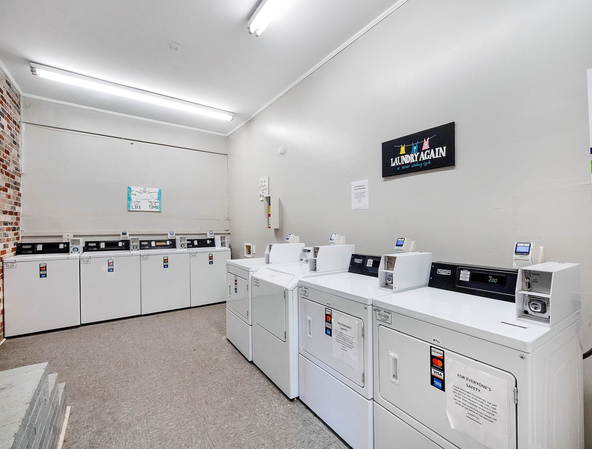 A laundry room with two rows of white commercial dryers along the walls, under bright overhead fluorescent lights.