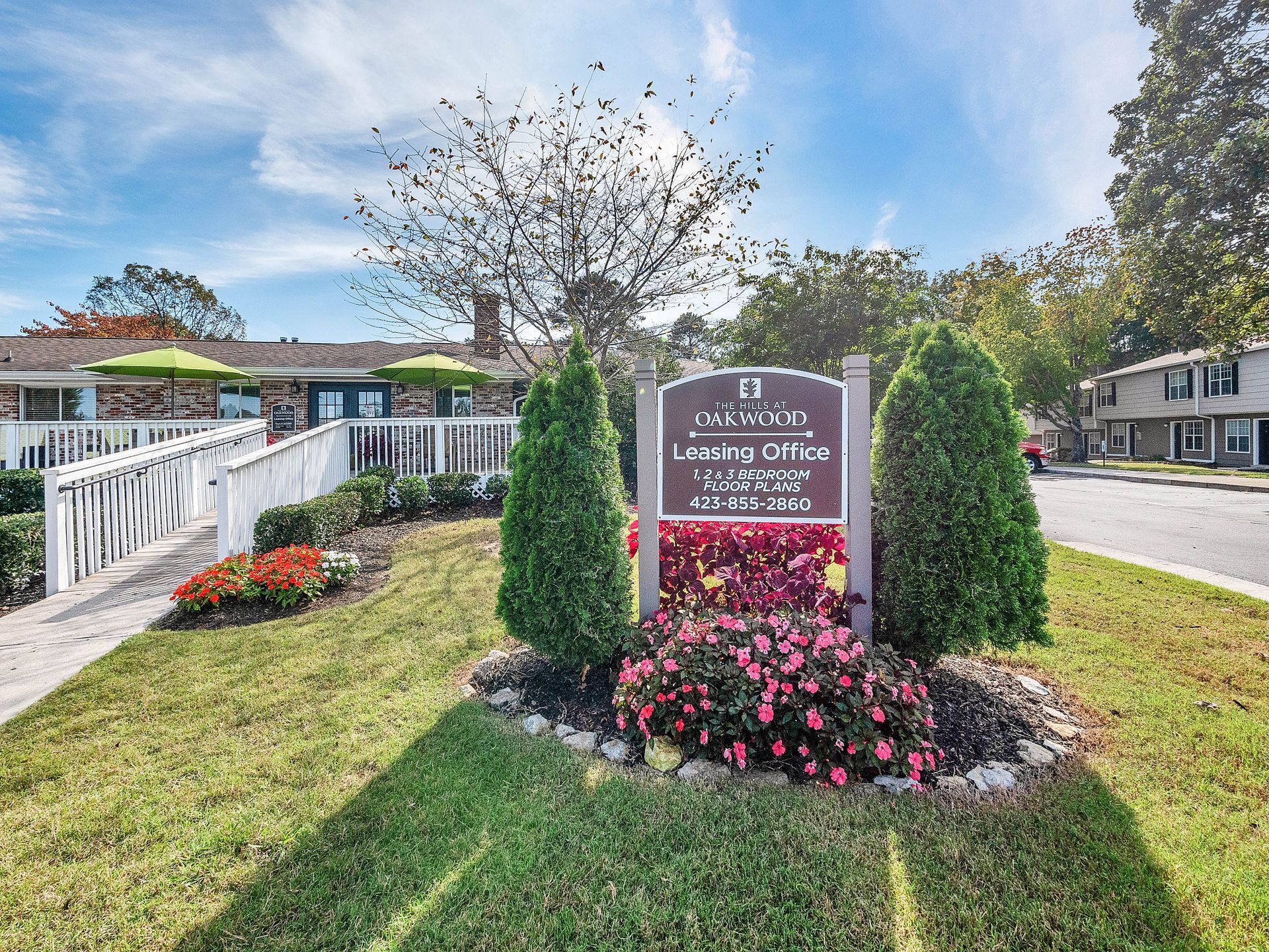 A sign for a leasing office stands in a garden bed with red flowers and two evergreen bushes, next to a white fence.