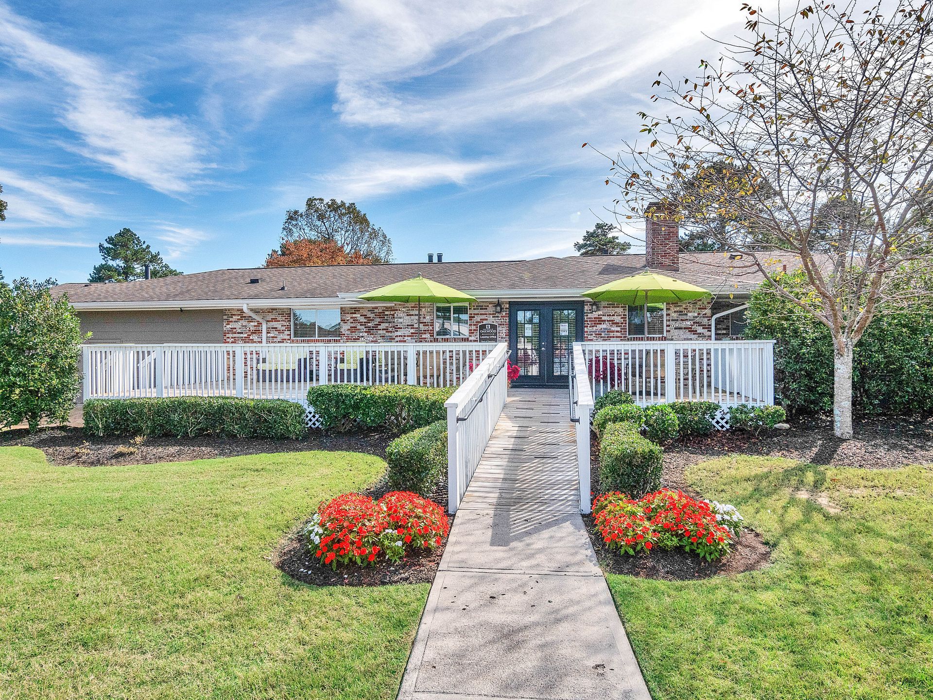 A sidewalk leads to a wide, white-railed deck with two green umbrellas in front of a brick one-story house.
