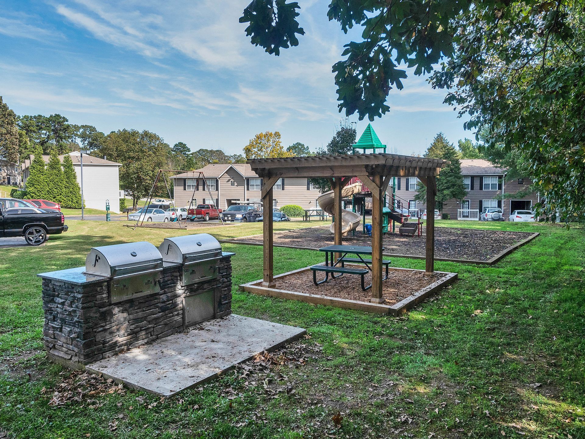 Outdoor community area with a double stainless steel grill, a wooden pergola over a picnic table, and apartment buildings.