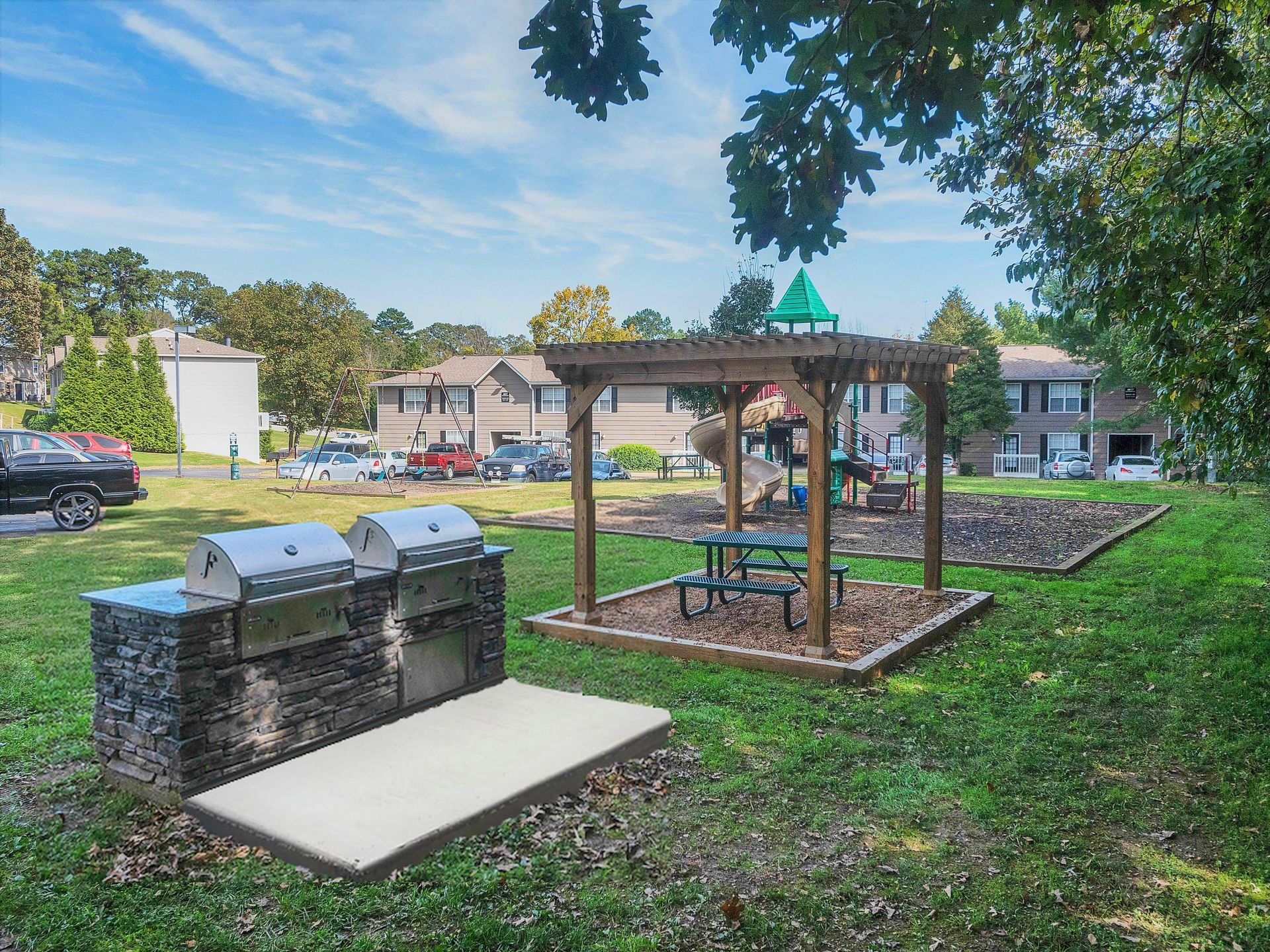 A park area with a double stainless steel stone grill, a wooden pergola with a picnic table, and a playground background.
