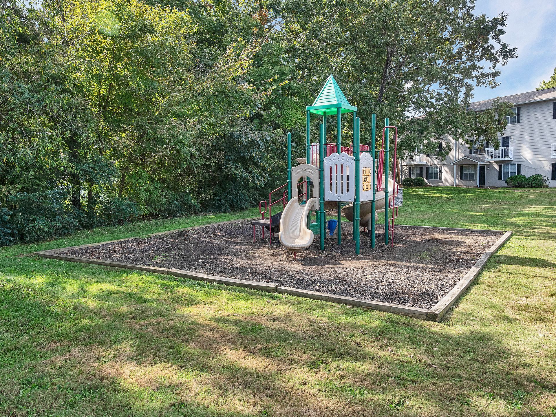 A small playground structure with a green roof, white panels, and a slide sits on a wood-mulch surface in a grassy yard.