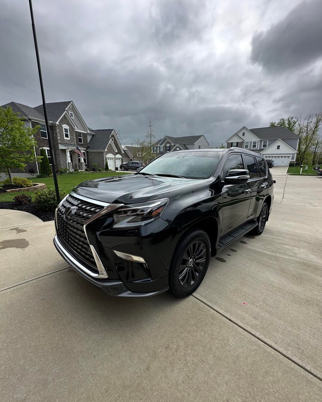 Black Lexus SUV parked on a driveway with houses in the background under a cloudy sky.