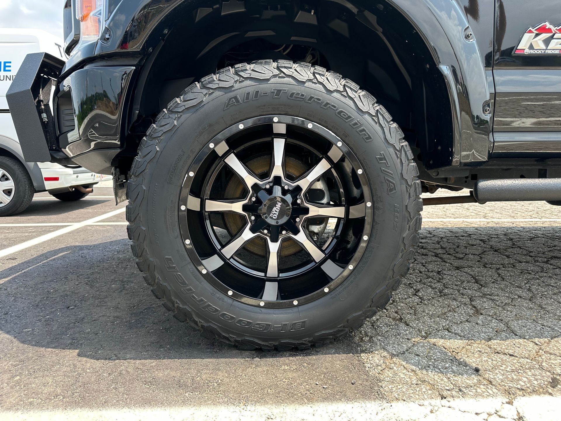 A black truck with a black and white tire is parked in a parking lot.