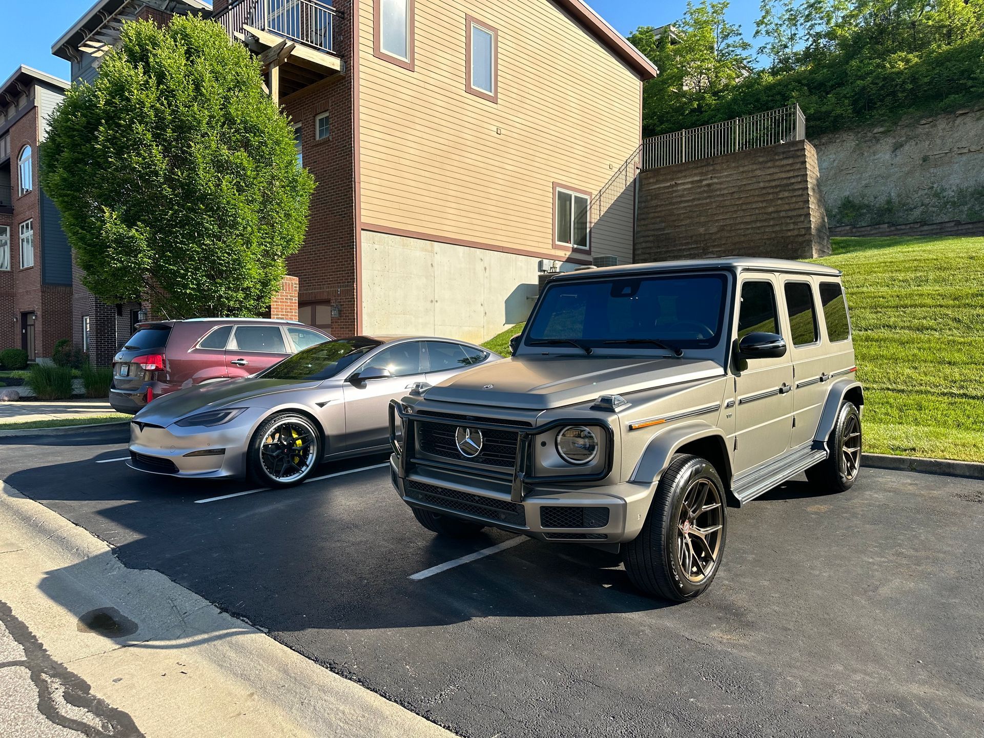A tan Mercedes-Benz G-Class SUV parked next to a silver Tesla sedan in a parking lot in front of a brick building.
