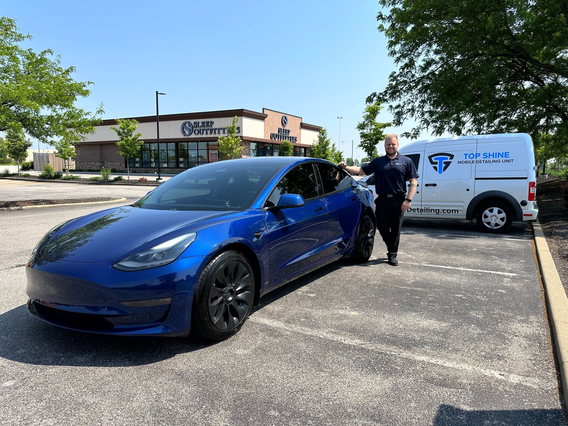 Blue Tesla parked by a white service van while a worker stands beside it in a parking lot.