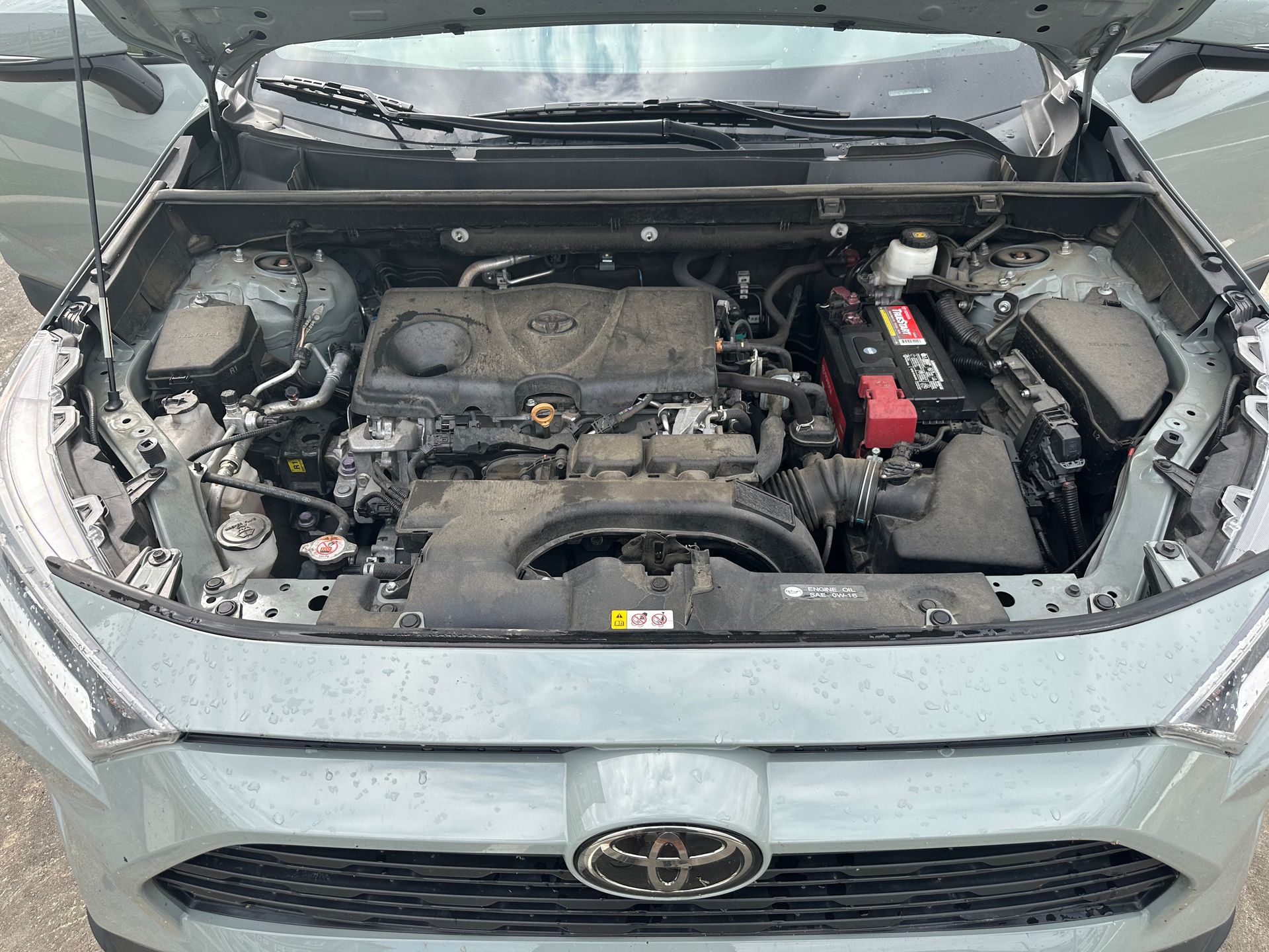 Open hood of a green Toyota RAV4, revealing its engine compartment. The engine is dusty, with various components visible.