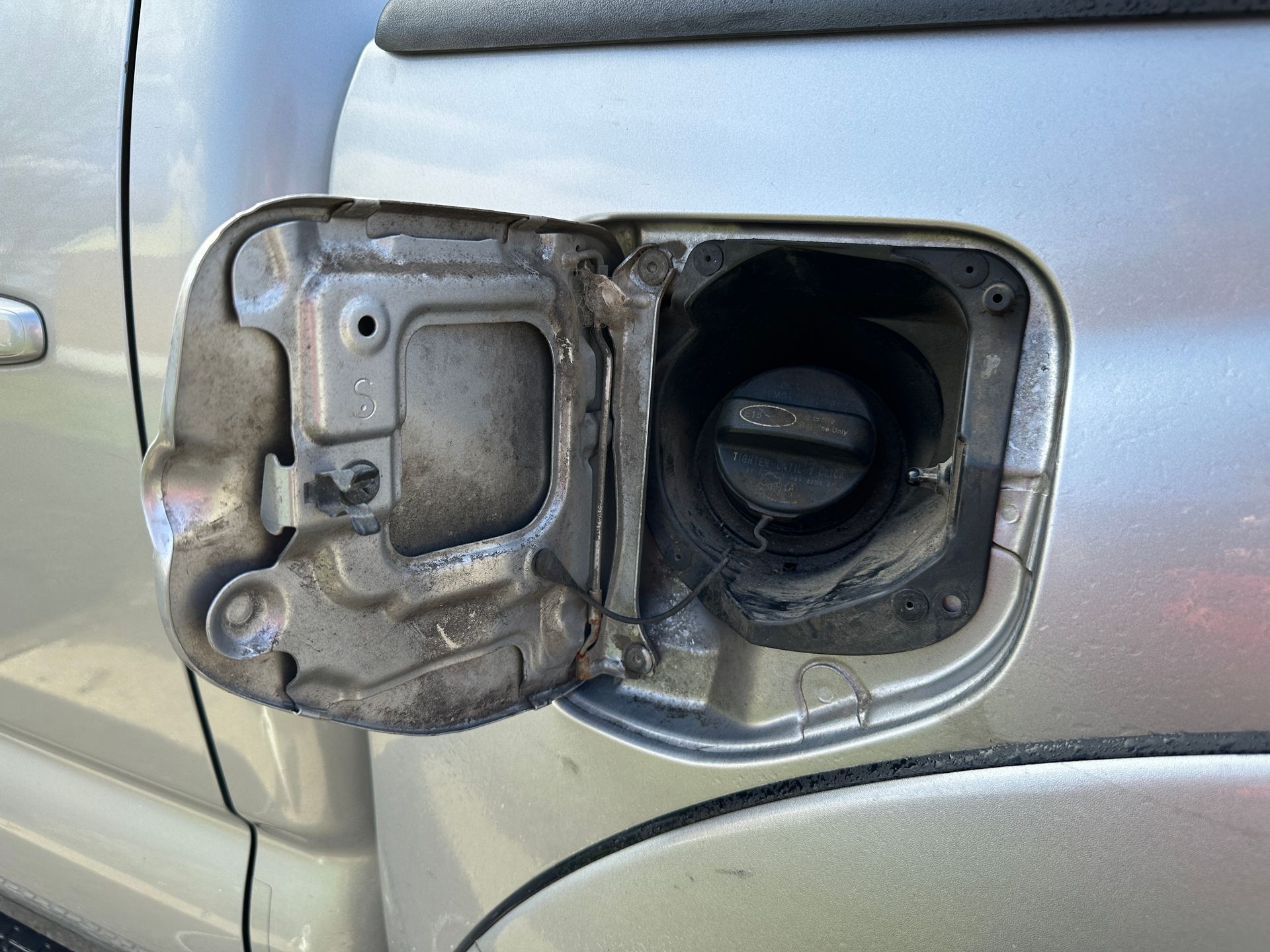 Open fuel door of a silver car, showing the fuel filler neck and cap, with dirt and wear present.